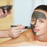 Beautician applying clay or carbon mask on face of young client in spa salon