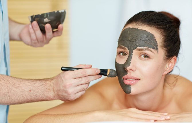 Beautician applying clay or carbon mask on face of young client in spa salon